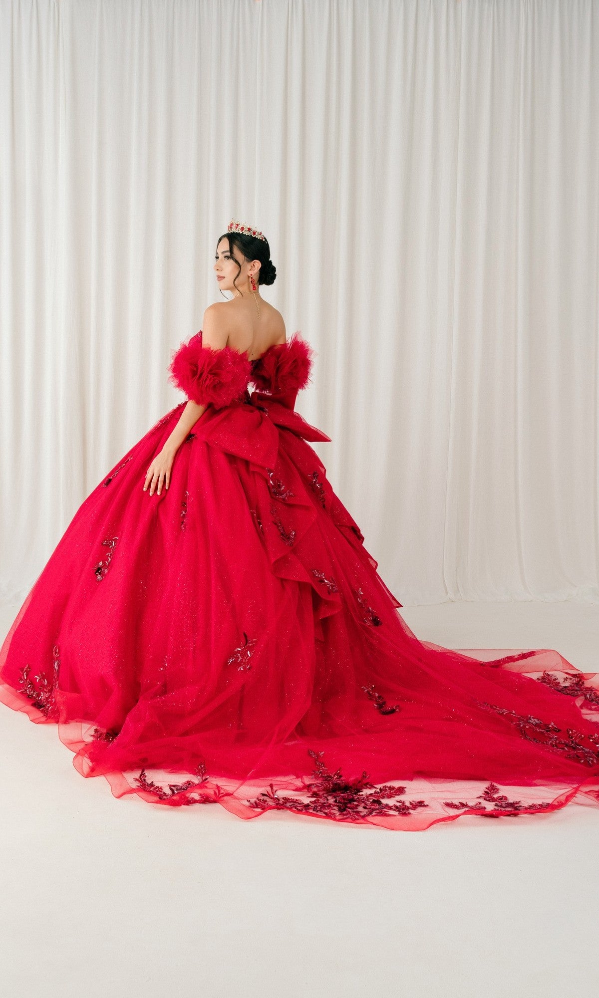 A woman faces away from the camera in front of white curtains, wearing the Quinceanera Dress 8380J by Cinderella Couture—an elaborate off-shoulder red ball gown with floral details, a corset bodice, and a long train.